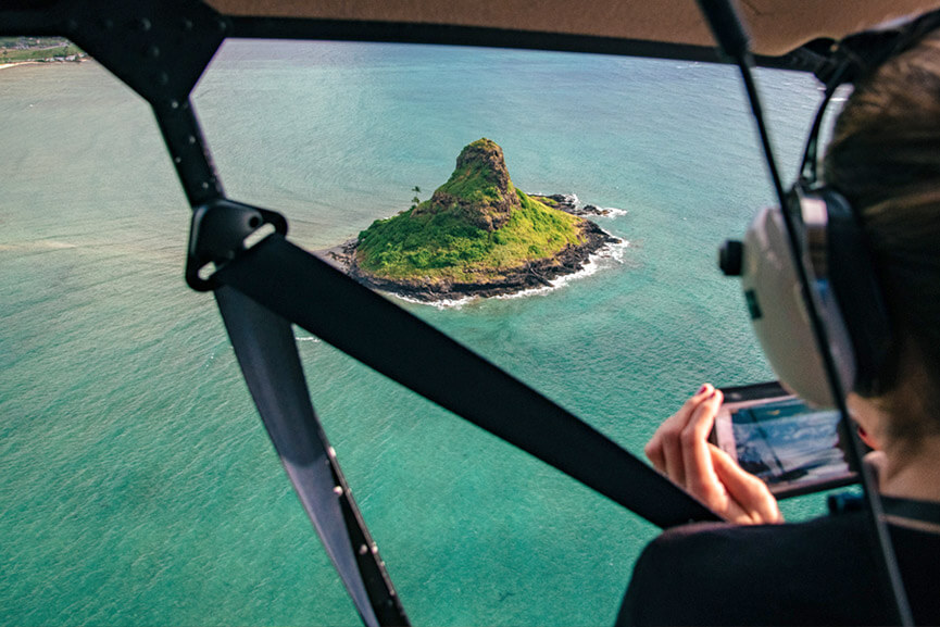 A person in a Rainbow Helicopter with doors off taking a photo of island and ocean below