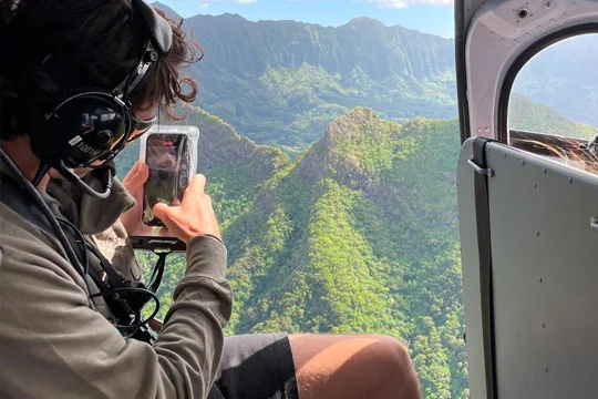 Young man taking picture with phone of epic green Hawaii landscape from a doors off Oahu helicopter tour.